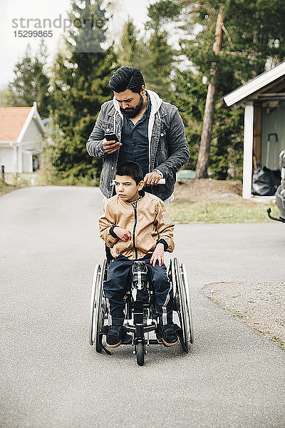Vater benutzt Smartphone  während er seinen autistischen Sohn im Rollstuhl schiebt