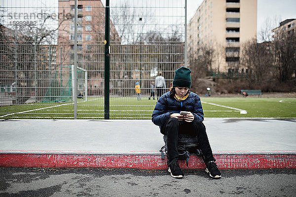 Ein Junge in voller Länge trägt warme Kleidung  während er mit seinem Handy gegen ein Fußballfeld in der Stadt telefoniert