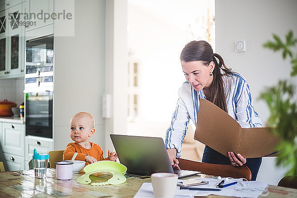 Weibliche Berufstätige hält Akte  während sie den Laptop ihrer Tochter auf dem Esstisch im Heimbüro benutzt