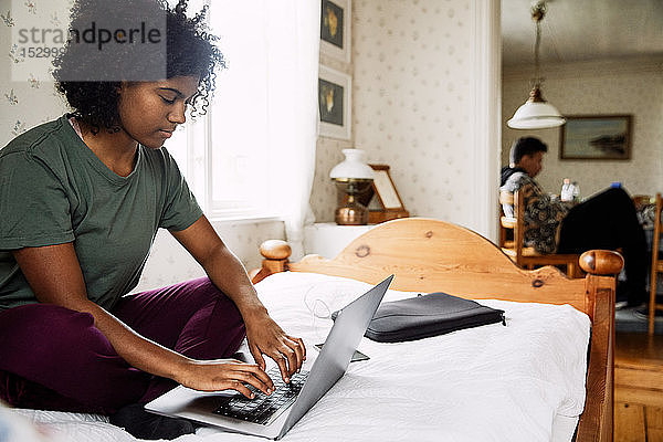 Junge Frau benutzt Laptop am Bett  während ein Freund zu Hause im Hintergrund sitzt