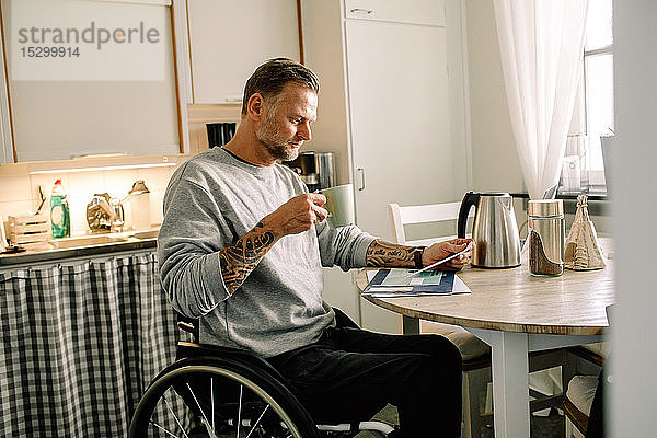 Tätowierter reifer Mann liest Post  während er zu Hause eine Kaffeetasse im Rollstuhl hält