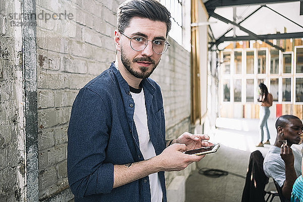 Porträt eines selbstbewussten jungen Geschäftsmannes  der mit einem Smartphone an einer Ziegelmauer an einem kreativen Arbeitsplatz steht