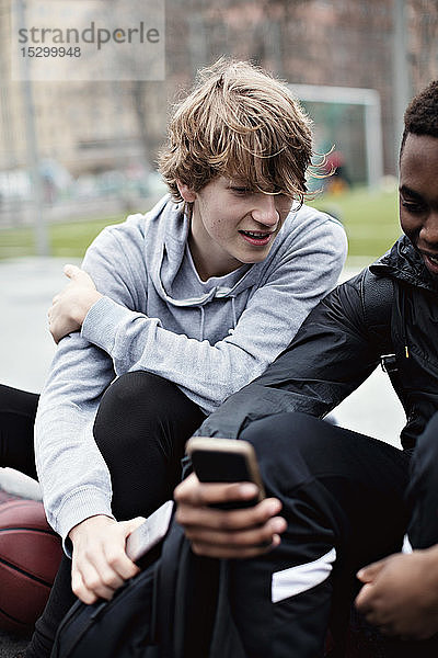 Teenager  der nach dem Basketballtraining auf dem Bürgersteig sitzend auf das Handy eines Freundes schaut