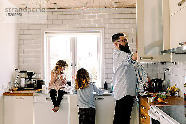 Mädchen sieht arbeitende Schwester an  während der Vater in der Küche arbeitet