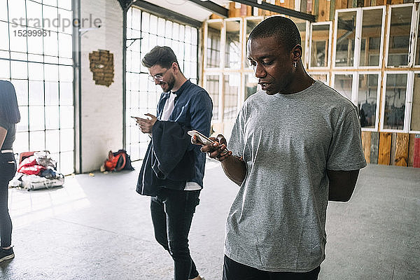 Selbstbewusster junger Geschäftsmann benutzt ein Smartphone  während er am kreativen Arbeitsplatz an einem Kollegen vorbeigeht