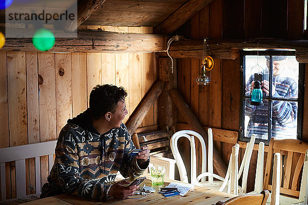 Mann fotografiert Freund beim Online-Shopping im Ferienhaus vom Fenster