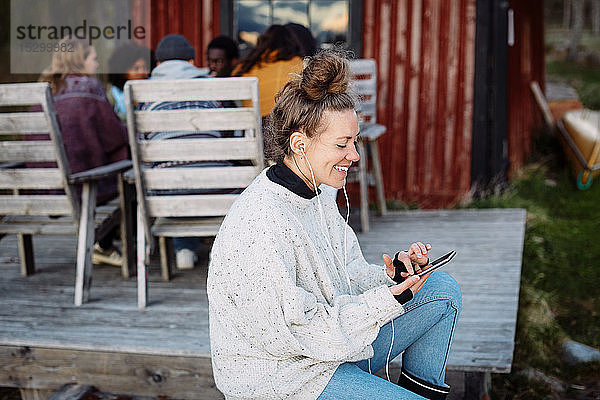 Lächelnde Frau mittleren Alters  die ein Smartphone benutzt  während Freunde im Hintergrund bei Sonnenuntergang sprechen