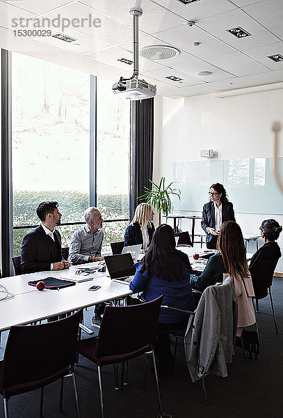 Geschäftsfrau interagiert mit einer Kollegin  die während einer Sitzung im Sitzungssaal mit Kollegen am Konferenztisch sitzt