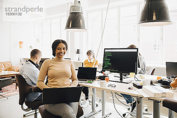 Porträt einer lächelnden Programmiererin  die einen Laptop benutzt  während ihre Kollegen im Hintergrund arbeiten