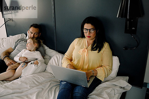 Hochwinkelansicht einer Frau am Laptop  während sie neben Mann und Tochter am Bett sitzt