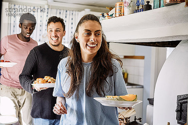 Lächelnde Freunde halten Essen in der Hand  während sie zu Hause in der Küche stehen