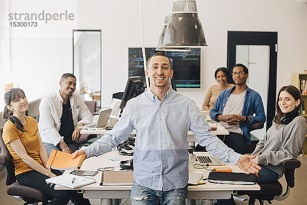 Porträt eines lächelnden Geschäftsmannes  der im Stehen mit Kollegen am Schreibtisch im Kreativbüro gestikuliert