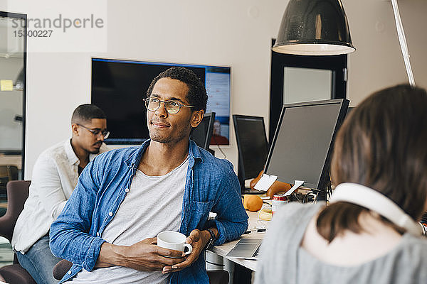 Nachdenklicher männlicher Computerprogrammierer schaut weg  während er neben Kollegen am Schreibtisch im Büro sitzt