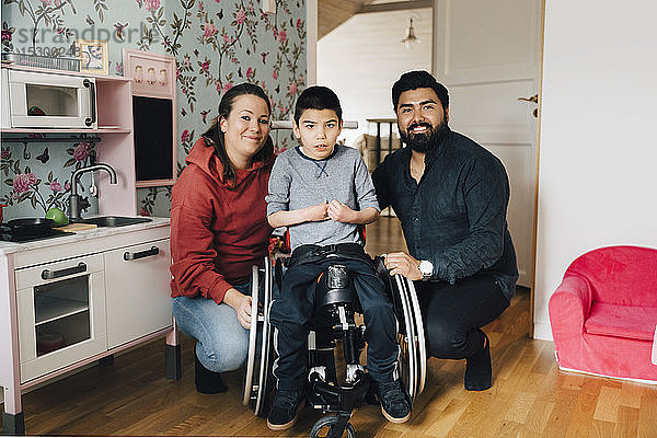 Porträt von lächelnden Eltern  die sich neben dem autistischen Sohn kauern  der zu Hause im Rollstuhl sitzt