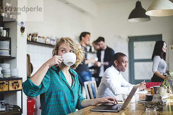 Junge Geschäftsfrau trinkt Kaffee  während Computerprogrammierer im Hintergrund im Büro arbeiten