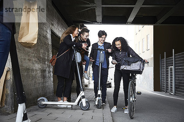 Glückliche männliche und weibliche Teenager-Freunde mit elektrischen Rollern schauen auf das Handy unter der Brücke