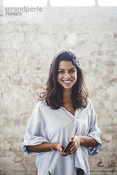 Porträt einer lächelnden jungen Computerprogrammiererin mit einem Smartphone in der Hand  die im Kreativbüro an einer Ziegelmauer steht