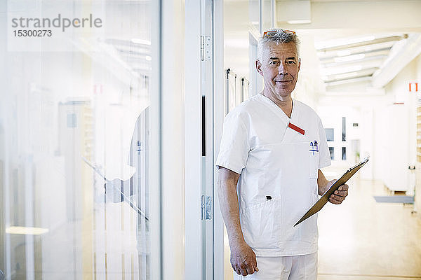 Porträt eines selbstbewussten leitenden Arztes  der ein Klemmbrett hält  während er im beleuchteten Korridor des Krankenhauses steht