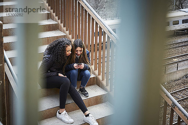 Teenager-Mädchen zeigt einer Freundin ein Handy  während sie auf einer Treppe sitzt
