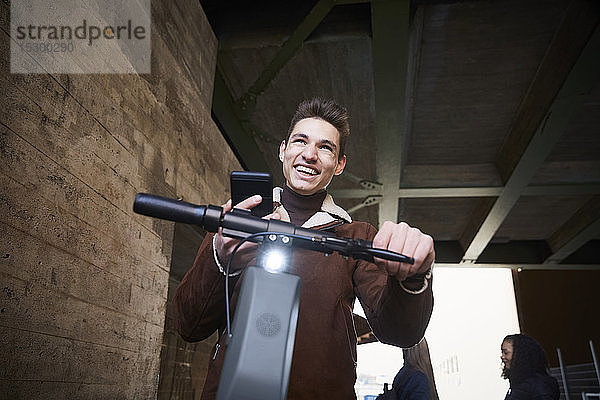 Fröhlicher Teenager mit elektrischem Schubsroller schaut weg  während er sein Mobiltelefon unter der Brücke hält