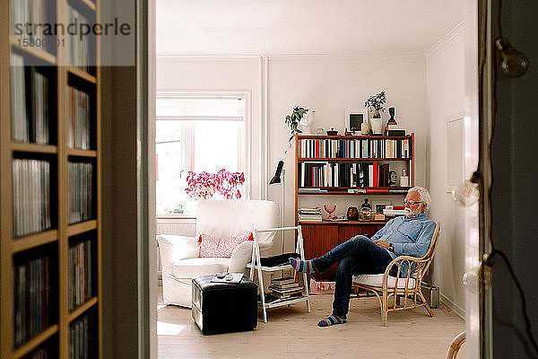 Älterer Mann sitzt in voller Länge auf einem Stuhl im Wohnzimmer von der Tür aus gesehen