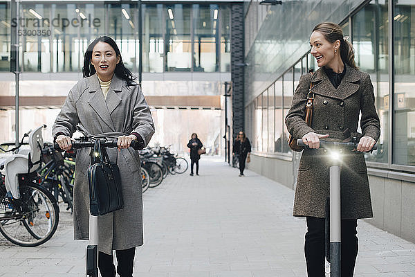 Lächelnde Kolleginnen auf Elektro-Schieberoller auf einem Fußweg durch ein Gebäude in der Stadt