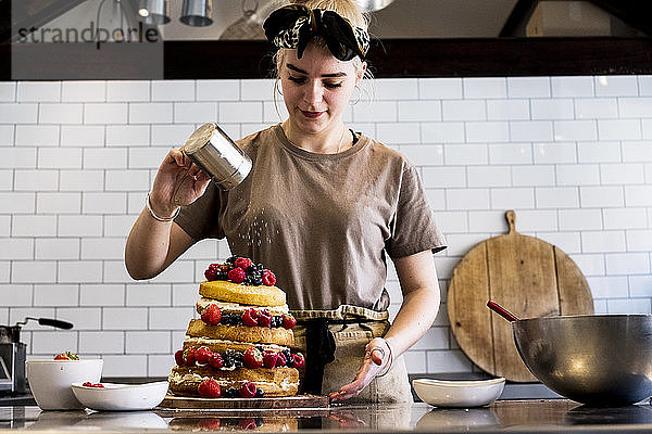 Ein Koch  der in einer Großküche arbeitet und Puderzucker über einen Schichtkuchen mit frischem Obst streut.