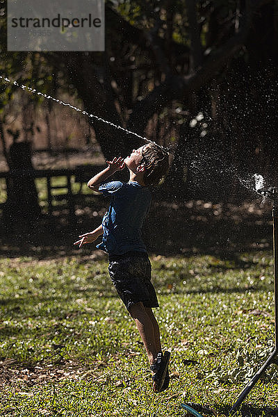 Unbekümmerter Junge springt in den Sprinkler im sonnigen Hinterhof