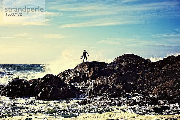 Silhouette eines männlichen Surfers  der die Wellen beobachtet  die an einem sonnigen Strand gegen die Felsen schlagen  Tofino  Kanada