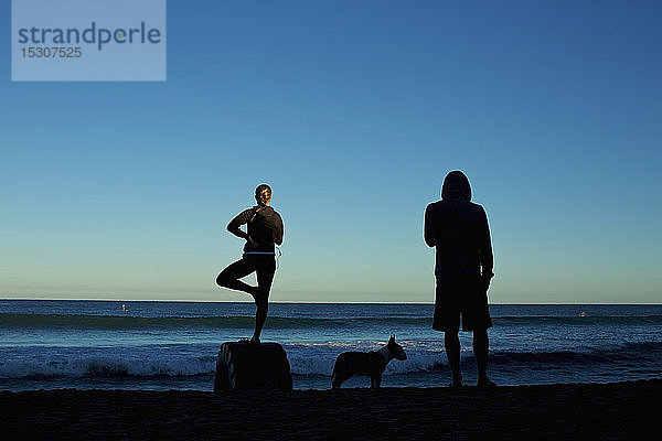 Silhouette junges Paar mit Hund übt Yoga auf Sonnenaufgang Strand