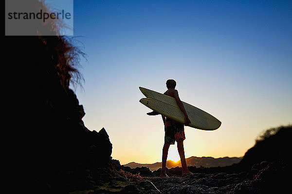 Silhouette Junge mit Surfbrett am idyllischen Strand bei Sonnenuntergang