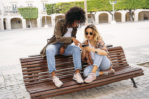 Multikulturelle glückliche Frauen  die sich unterhalten und ein Smartphone benutzen  auf einer Bank sitzend