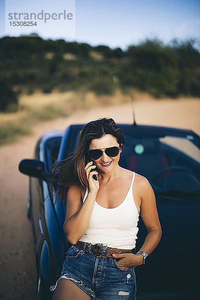 Sexy Frau  die sich an ein Cabrio lehnt und ein Smartphone benutzt