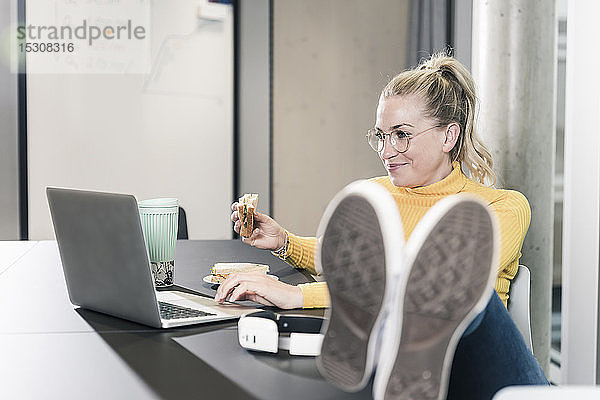 Lächelnde  ungezwungene Geschäftsfrau  die im Büro am Tisch sitzt  ihren Laptop benutzt und ein Sandwich isst