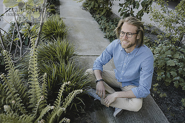Lächelnder junger Mann sitzt auf dem Bürgersteig umgeben von Pflanzen