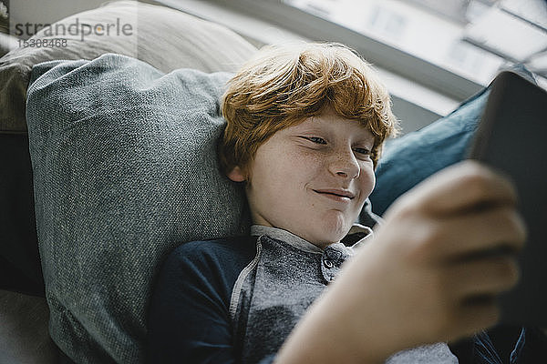 Porträt eines lächelnden rothaarigen Jungen  der mit Hilfe eines digitalen Tablets auf der Couch liegt