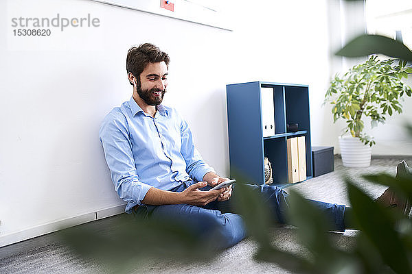 Lächelnder Geschäftsmann  der auf dem Boden sitzt und sein Smartphone benutzt