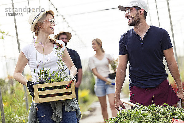Lächelnder Mann und Frau beim Transport von Pflanzen mit Schubkarre und Holzkiste in einem Gewächshaus