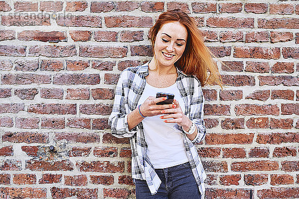 Junge Frau lehnt in der Stadt an Ziegelmauer  während sie ein Smartphone benutzt
