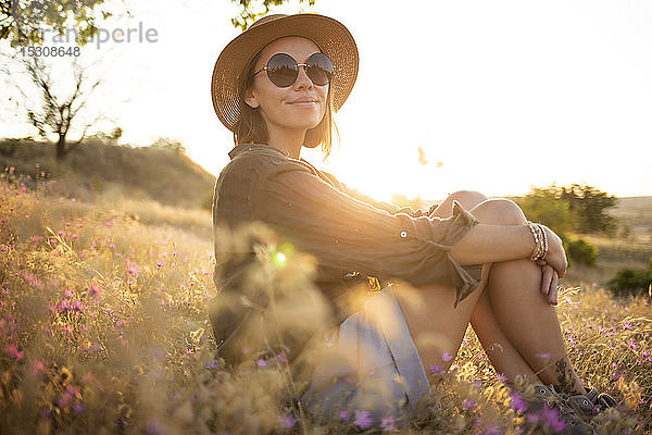 Junge Frau trägt Sonnenhut und sitzt bei Sonnenuntergang auf der Wiese