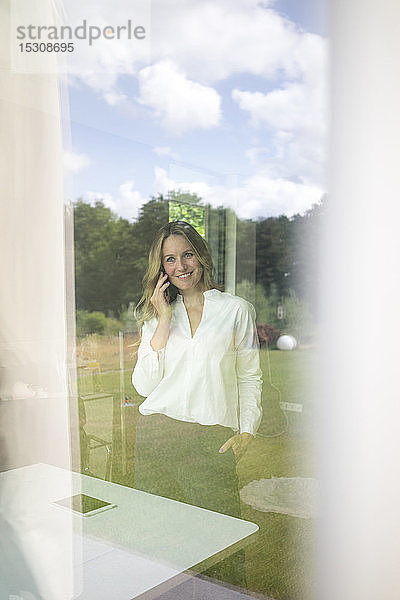 Lächelnde Geschäftsfrau telefoniert zu Hause hinter einer Fensterscheibe