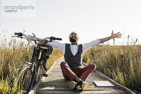 Glücklicher  gut gekleideter Mann sitzt auf einem Holzsteg auf dem Land neben einem Fahrrad