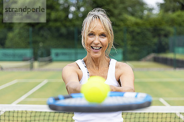 Porträt einer lachenden  reifen Frau  die im Tennisclub einen Tennisschläger mit einem Ball in der Hand hält