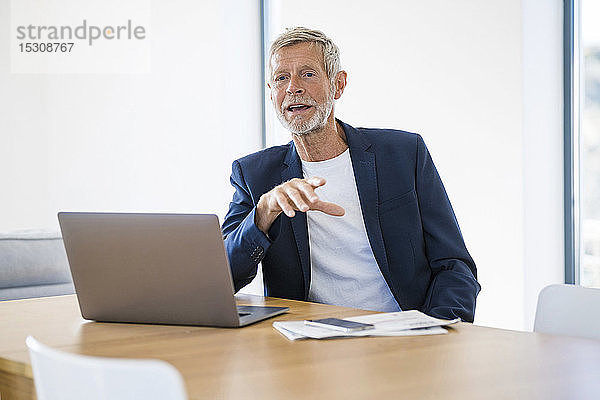Leitender Geschäftsmann mit Laptop sitzt zu Hause am Schreibtisch und unterhält sich