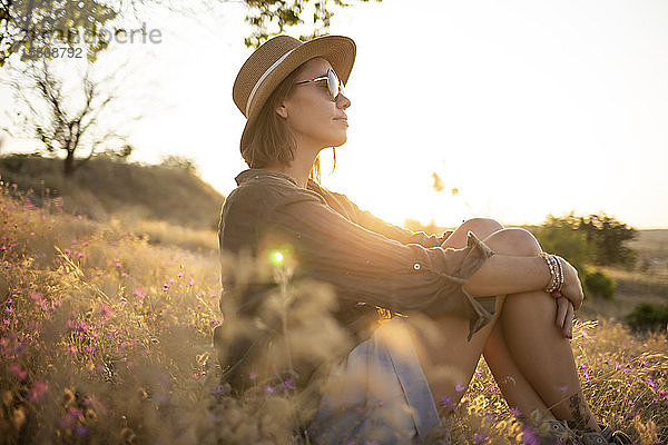Junge Frau mit Sonnenhut  die bei Sonnenuntergang auf einer Wiese sitzt