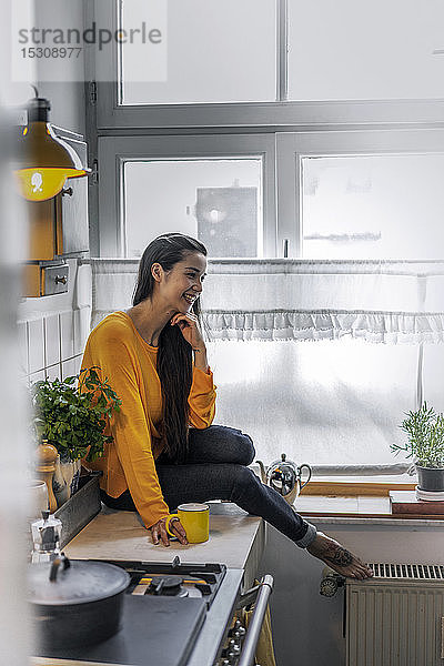 Glückliche junge Frau sitzt zu Hause auf dem Küchentisch
