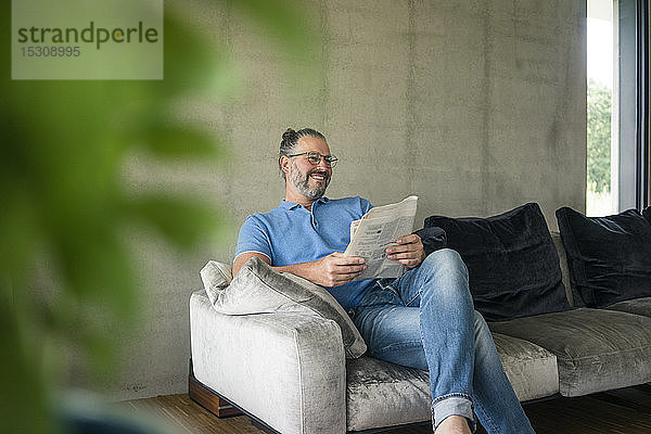 Lächelnder reifer Mann sitzt zu Hause auf der Couch und liest Zeitung