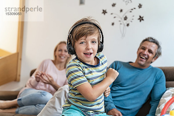 Porträt eines Jungen  der zu Hause auf der Couch mit Kopfhörern Musik hört  mit den Eltern im Hintergrund
