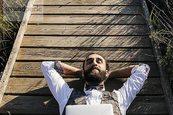 Gut gekleideter Mann mit Laptop  der auf einem Holzsteg auf dem Land liegt
