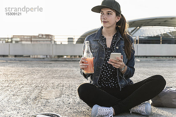 Junge Frau mit Getränk und Mobiltelefon bei Sonnenuntergang auf dem Parkdeck sitzend